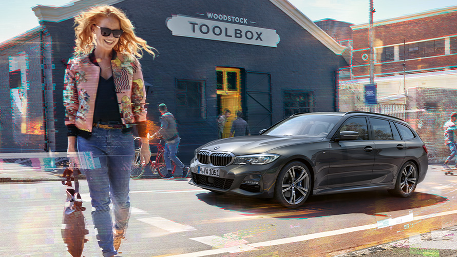 BMW 3 SeriesTouring, (G21) three-quarter front shot with woman in front of a woodworks-toolshop. Woman crosses the street in front of a BMW 3 Series Touring (G21) in front of a woodworks-toolshop.