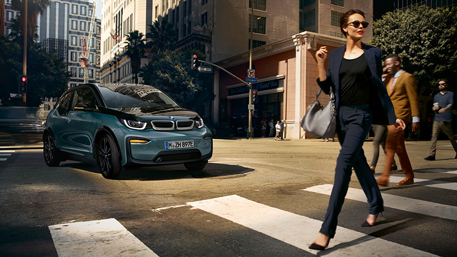 BMW i3 I01 three-quarter front view with woman walking accross zebra crossing BMW i3 I01 Blue Ridge Mountain metallic with Frozen Grey metallic accent three-quarter front view with woman walking accross zebra crossing