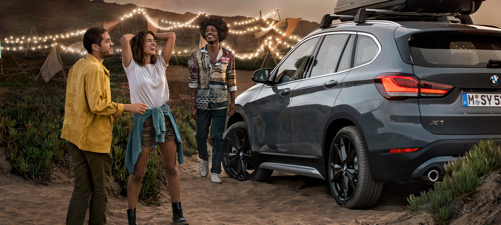 Cheering trio in BMW X1 with BMW offers summer Happy trio in BMW X1 Mineral Grey equipped with BMW offers summer three quarter rear view at the beach BMW equipment