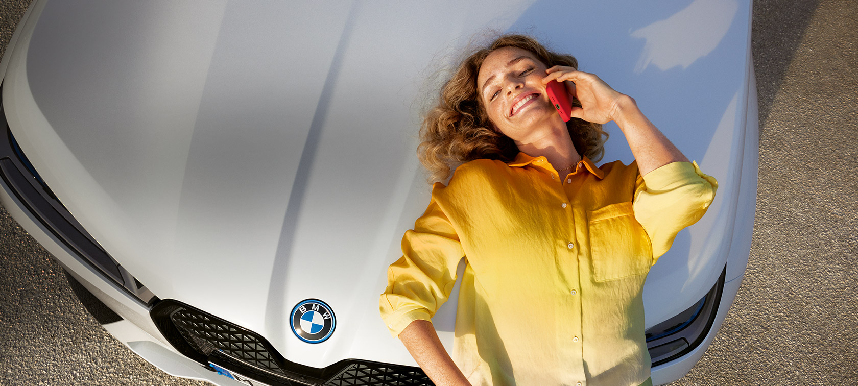 Relax We Care Proactive Care, Check-up & Maintenance Proactive Care, Check-up & Maintenance Relax We Care Woman with yellow shirt and smartphone in hand leaning on white BMW bonnet